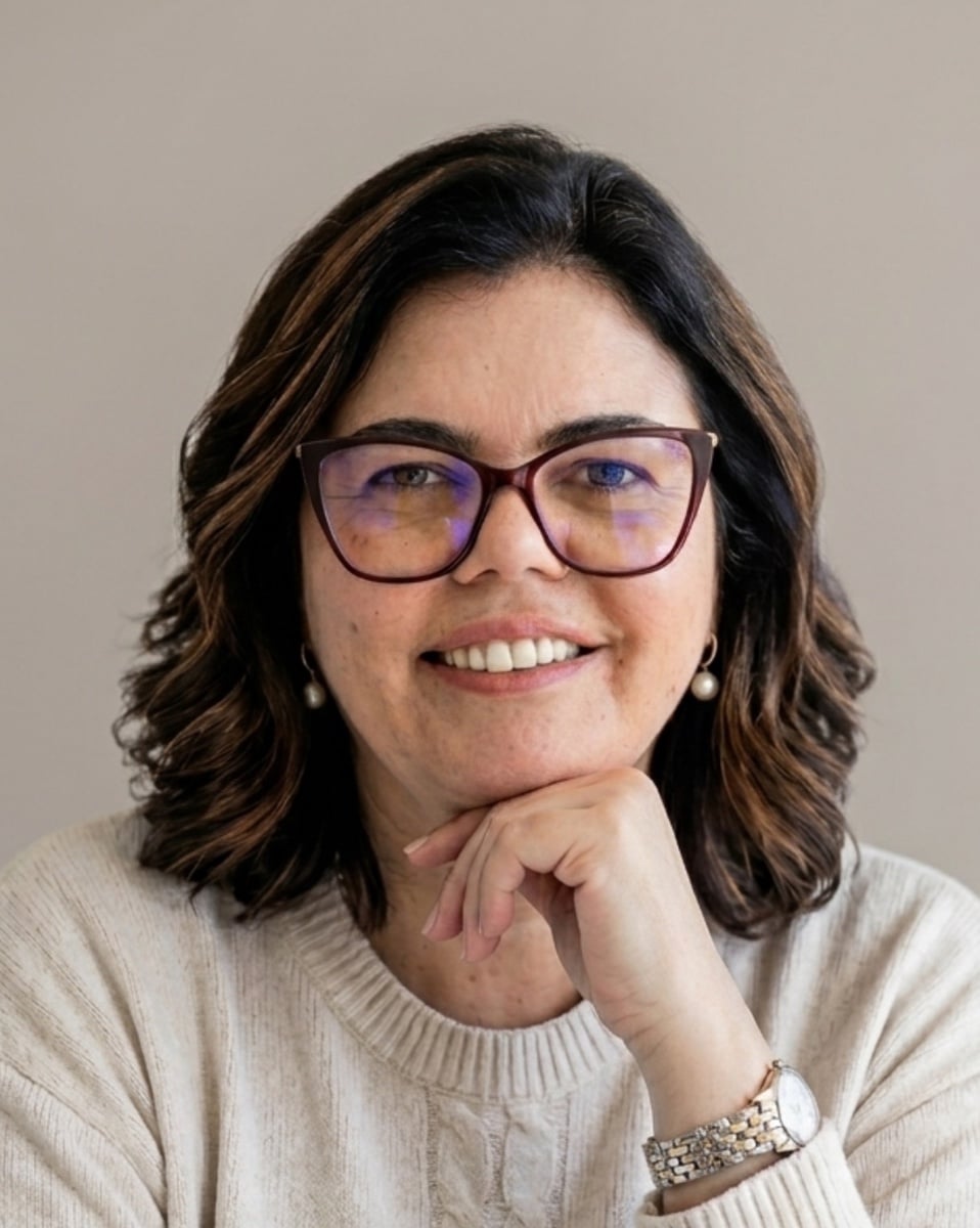 Cláudia Goulart, arquiteta e gestora de tráfego, sorri com a mão no queixo, de camisola bege, num estúdio de luz suave.